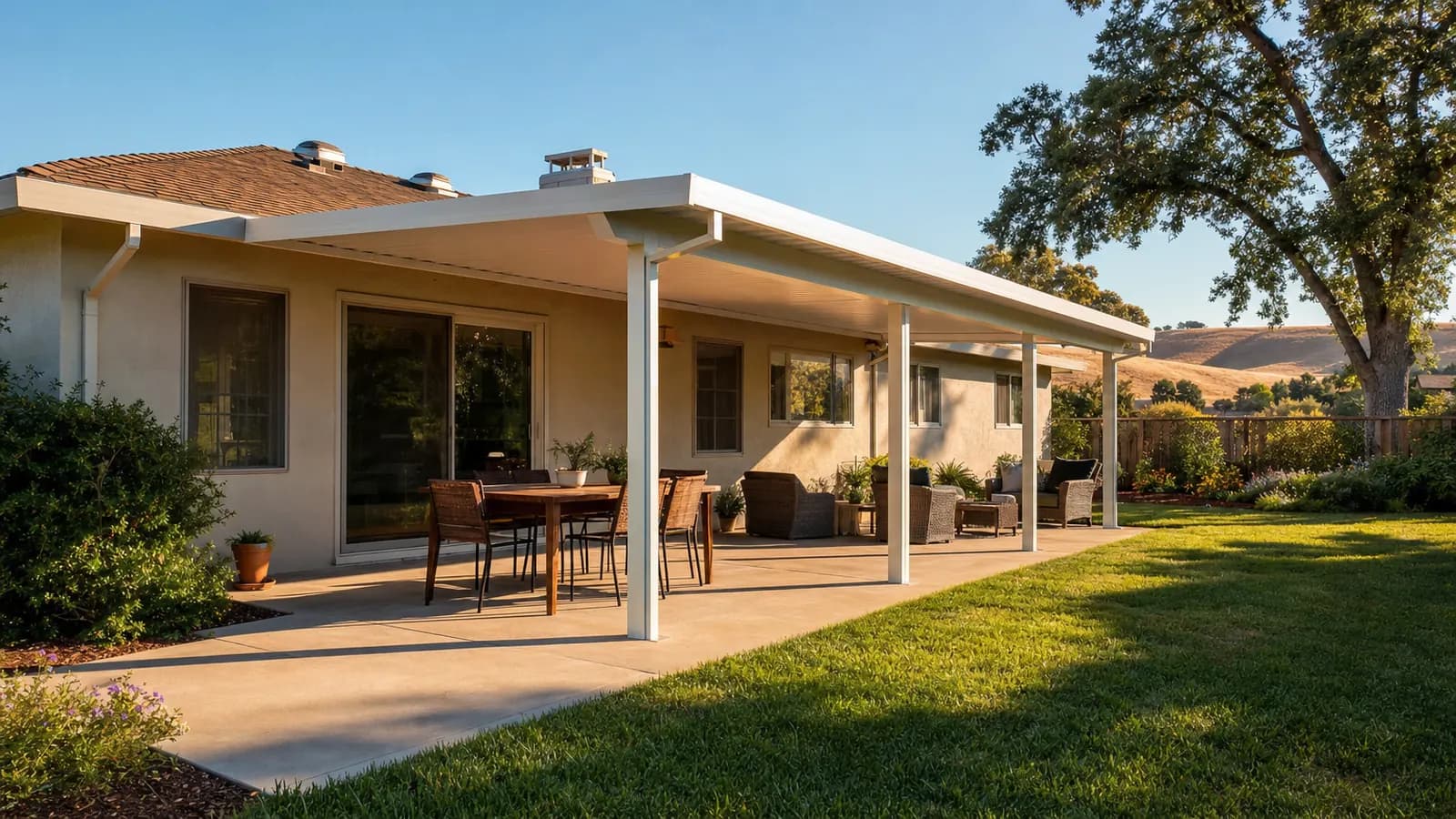 White solid aluminum patio cover attached to a single-story stucco ranch home, shading a concrete patio with a dining table, with a valley oak and golden hills behind the lawn